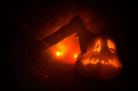 Scary orange pumpkin with carved eyes and a smile with burning candles and an ax on a dark background with fire sky.の写真素材
