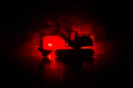 silhouette tracked excavator on dark foggy red orange sky background with toy carの写真素材