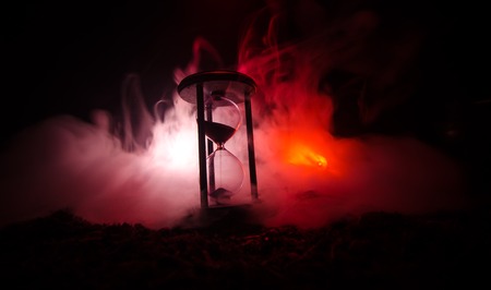 Time concept. Silhouette of Hourglass clock and old vintage wood clock with arrow and smoke on dark background with hot yellow orange red blue cold back lighting, or symbols of time with copy spaceの写真素材