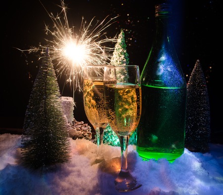 New Years Eve celebration background with pair of flutes and bottle of champagne with christmas tree on snow on dark background. Selective focusの写真素材