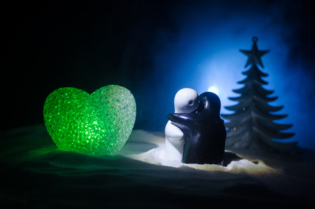 Love New Year concept. Girl and boy ceramic figures hugs each other, standing on the white snow and colored heart and Christmas tree on blurred dark background. Empty space. Hugging sculptures.の写真素材