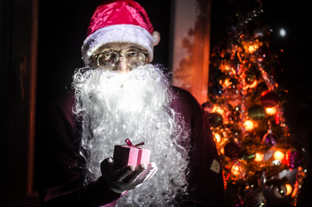 Santa Claus holding gift with festive holiday background. Santa Claus holding and offering a gift box on his hand. Selective focus. Dark toned backgroundの写真素材