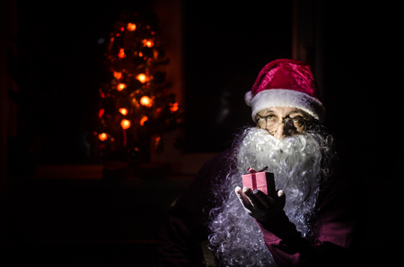 Santa Claus holding gift with festive holiday background. Santa Claus holding and offering a gift box on his hand. Selective focus. Dark toned backgroundの写真素材