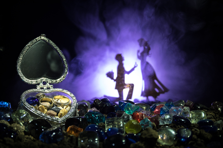 Beautiful opened old vintage silver heart-shaped casket with wedding rings with silhouette of young man staying on the knee and making proposal for his lovely girl. Floral dark foggy fairy background.の写真素材