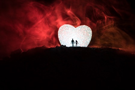 Silhouette of happy couple standing behind big shaped heart symbol on mountain at night. Big heart like moon glowing at foggy sky. Valentine`s day decor photo. Toned backgroundの写真素材