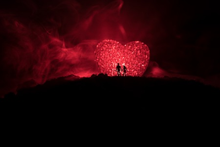 Silhouette of happy couple standing behind big shaped heart symbol on mountain at night. Big heart like moon glowing at foggy sky. Valentine`s day decor photo. Toned backgroundの写真素材