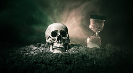 Skull and vintage hourglass on dark toned foggy background under beam of light. Horror concept. Empty space.の写真素材