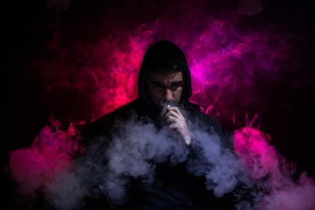 Vaping man holding a mod. A cloud of vapor. dark foggy toned background of clouds of smoke. Vaping...