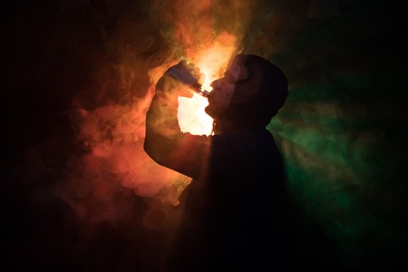 Vaping man holding a mod. A cloud of vapor. dark foggy toned background of clouds of smoke. Vaping an electronic cigarette with a lot of smoke. Vape concept. Selective focusの写真素材