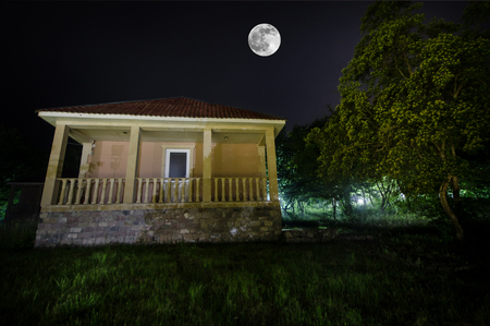 Mountain night landscape of building at forest in foggy night with moon. Green meadow, big trees and abandoned house at nightの写真素材