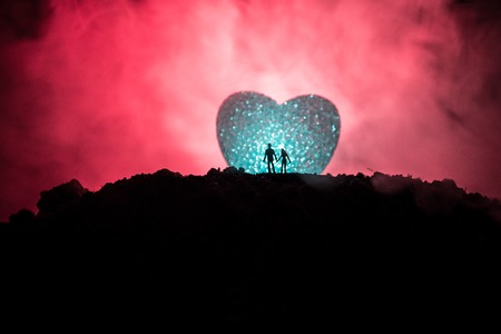 Silhouette of happy couple standing behind big shaped heart symbol on mountain at night. Big heart like moon glowing at foggy sky. Valentine`s day decor photo. Toned backgroundの写真素材