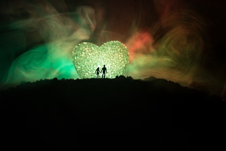 Silhouette of happy couple standing behind big shaped heart symbol on mountain at night. Big heart like moon glowing at foggy sky. Valentine`s day decor photo. Toned backgroundの写真素材
