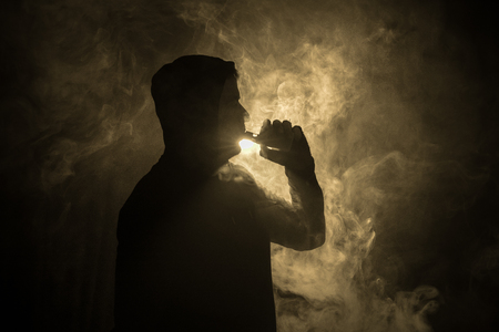 Vaping man holding a mod. A cloud of vapor. dark foggy toned background of clouds of smoke. Vaping an electronic cigarette with a lot of smoke. Vape concept. Selective focusの写真素材