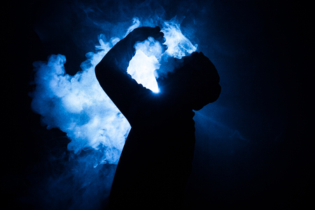 Vaping man holding a mod. A cloud of vapor. dark foggy toned background of clouds of smoke. Vaping an electronic cigarette with a lot of smoke. Vape concept. Selective focusの写真素材