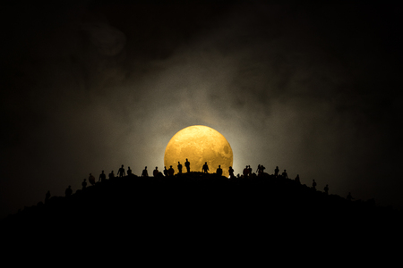 Scary view crowd of zombies on hill with spooky cloudy sky with fog and rising full moon. Silhouette group of zombie walking under full moon. Halloween concept.の写真素材
