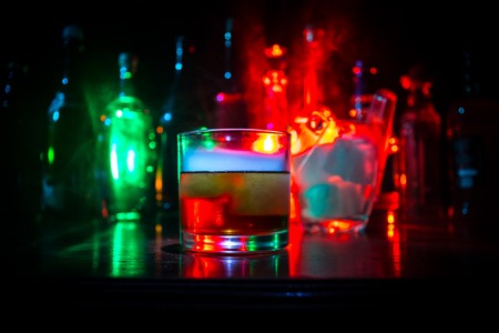 Glass of whisky on wooden bar closeup with bottles blurred view on dark background with light and...