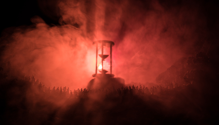 Silhouette of a large crowd of people in forest at night standing against a big hourglass with toned light beams on foggy background. Time concept. Hourglass measuring the passing timeの写真素材