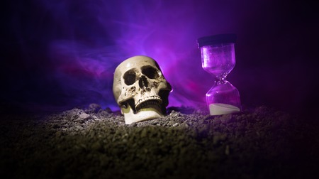 Skull and vintage hourglass on dark toned foggy background under beam of light. Horror concept. Empty space.の写真素材