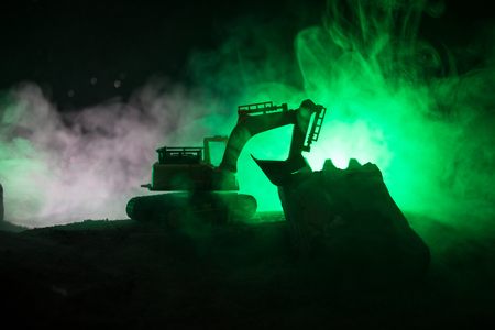 Construction site on a city street. A yellow digger excavator parked during the night on a construction site. Industrial concept table decoration on dark foggy toned background. Selective focusの写真素材