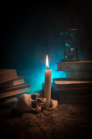 Wizard's Desk. A desk lit by candle light. A human skull, old books on sand surface. Halloween still-life background with a different elements on dark toned foggy background. Selective focusの写真素材