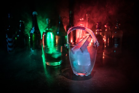 Glass ice bucket on blurred alcohol bottles background with lights and smoke. Club drinks concept....