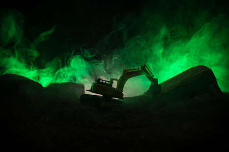 Construction site on a city street. A yellow digger excavator parked during the night on a construction site. Industrial concept table decoration on dark foggy toned background. Selective focusの写真素材