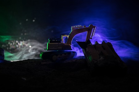Construction site on a city street. A yellow digger excavator parked during the night on a construction site. Industrial concept table decoration on dark foggy toned background. Selective focusの写真素材