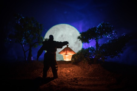Man in raincoat coming from dark forest with glowing lantern in his hand concept. Silhouette of a horror killer with lamp. Decorationの写真素材