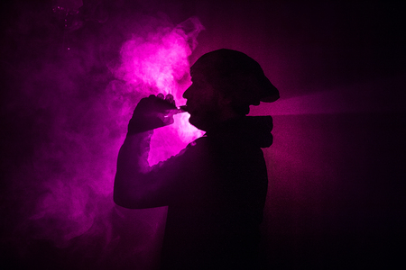 Vaping man holding a mod. A cloud of vapor. dark foggy toned background of clouds of smoke. Vaping...