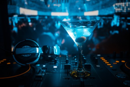 Glass with martini with olive inside on dj controller in night club. Dj Console with club drink at music party in nightclub with disco lights.の写真素材