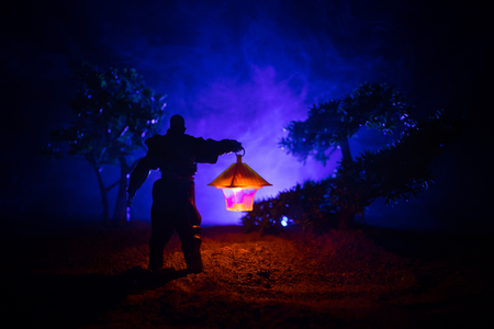 Man in raincoat coming from dark forest with glowing lantern in his hand concept. Silhouette of a horror killer with lamp. Decorationの写真素材