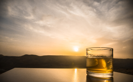 A glass of whiskey with ice on a sunset background or shot of whiskey at sunset dramatic sky on mountain landscape background. Selective focusの写真素材