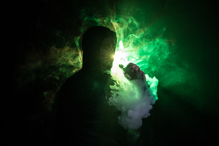 Vaping man holding a mod. A cloud of vapor. dark foggy toned background of clouds of smoke. Vaping an electronic cigarette with a lot of smoke. Vape concept. Selective focusの写真素材