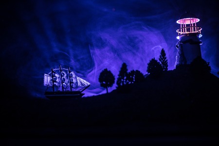 Old wooden warship sailing by night close to lighthouse or lighthouse and sailboat. Dark foggy background. Selective focus. Decorated photoの写真素材