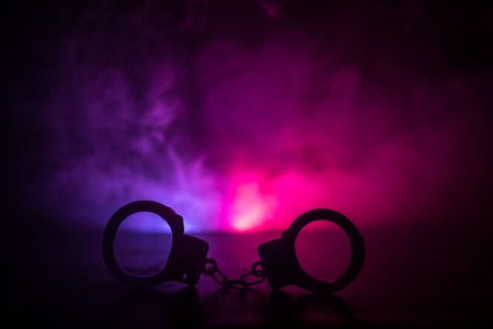 Police raid at night and you are under arrest concept. Silhouette of handcuffs with police car on backside. Image with the flashing red and blue police lights at foggy background. Slider shotの写真素材