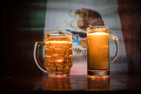 Creative concept. Beer glasses on table at dark toned foggy background with blurred view of flag of Mexico. Support your country with beer concept. Selective focusの写真素材