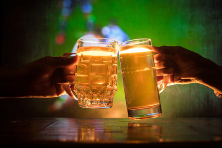 Two friends toasting (clinking) with glasses of light beer at the pub. Beautiful background with blurred view of playing game at the stadium. Selective focusの写真素材