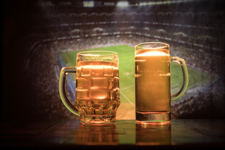 Creative concept. Beer glasses on table at dark toned foggy background with blurred view of playing game at the stadium. Selective focusの写真素材