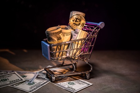 Close up view of little shopping trolley with a lot of money inside and Bitcoin (new virtual money psyhical version) coin. Selective focus. Digital money and Bitcoin concept.の写真素材