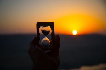 Life time passing concept. Hand holding hourglass with sunset sky background. Selective focusの写真素材