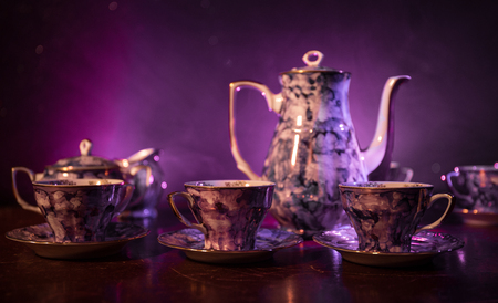 Coffee or tea ceremony conceptual theme. Old vintage ceramic tea or coffee pot with cups jug and sugar cup on dark toned background with light and smoke. Empty space for text. Selective focusの写真素材