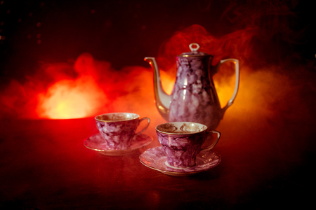 Coffee or tea ceremony conceptual theme. Old vintage ceramic tea or coffee pot with cups on dark toned background with light and smoke. Empty space for text. Selective focusの写真素材