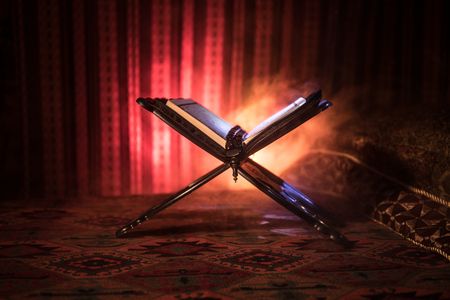 Open holy book of Muslims on stand on eastern carpet with dark toned foggy background. Muslim religion concept. Selective focus. Indoor shot.の写真素材