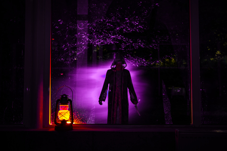 Horror Halloween concept. Burning old oil lamp standing on window with silhouette of an unknown shadow figure with backlight. Night scenery of a nightmare scene. Selective focus.の写真素材