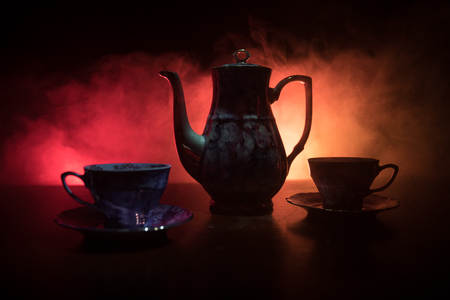 Coffee or tea ceremony conceptual theme. Old vintage ceramic tea or coffee pot with cups on dark toned background with light and smoke. Empty space for text. Selective focusの写真素材