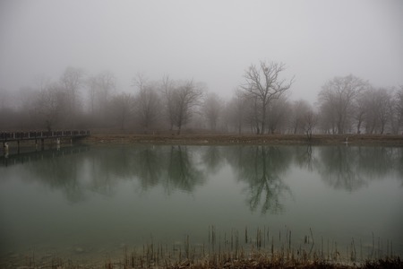 Amazing landscape of bridge reflect on surface water of lake, fog evaporate from pond make romantic scene or Beautiful bridge on lake with trees at fog. Iron bridge over lake in misty morning.の写真素材