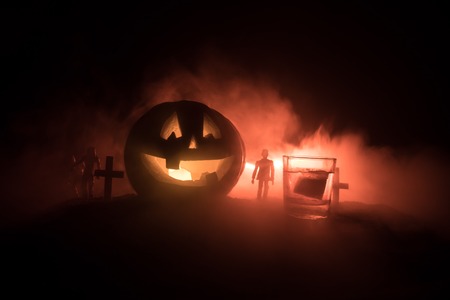 Halloween pumpkin with carved face and glass of whiskey with ice on a dark toned foggy background...