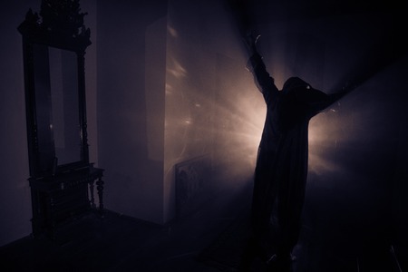 Horror silhouette of ghost inside dark room with mirror. Scary halloween concept. Silhouette of witch inside haunted house with fog and light on background. Selective focusの写真素材