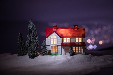 Christmas and New Year miniature house in the snow at night with fir tree. Little toy house on snow with tree and bokeh city lights on background. Christmas decorations. Holiday concept.の写真素材