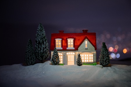 Christmas and New Year miniature house in the snow at night with fir tree. Little toy house on snow with tree and bokeh city lights on background. Christmas decorations. Holiday concept.の写真素材